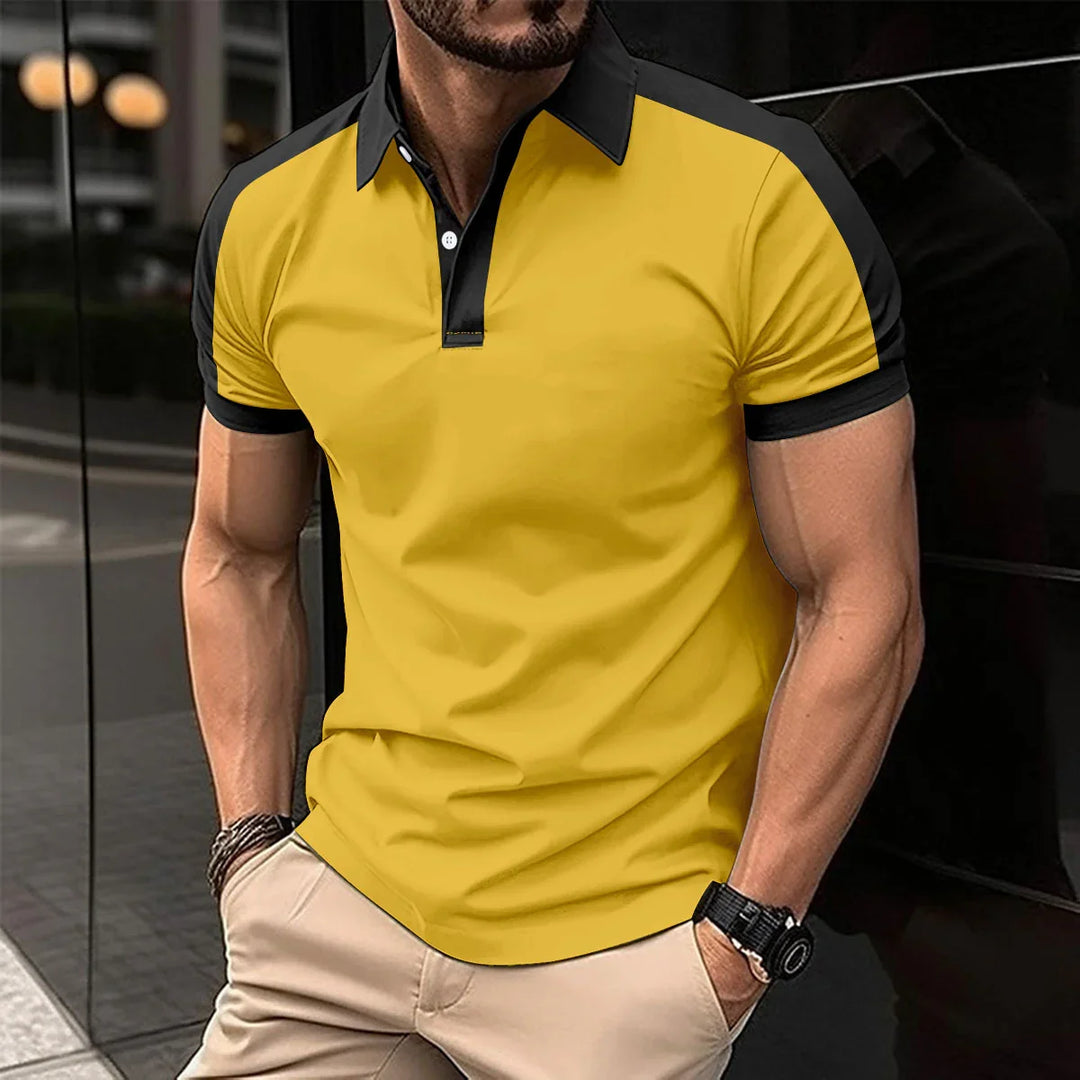 Man wearing yellow and black polo shirt with beige pants, standing against glass wall