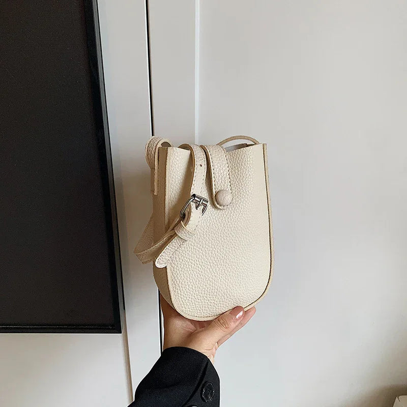 Hand holding a small textured beige leather crossbody bag with silver buckle against a white wall