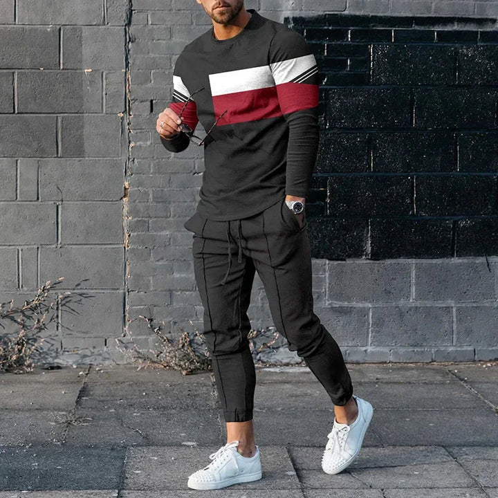Man wearing black color-block sweater with white and red stripes, black jogger pants, and white sneakers standing against a gray brick wall