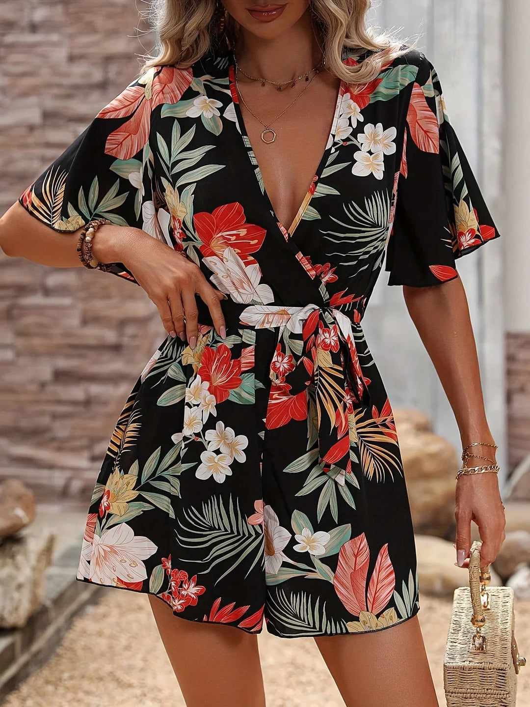 Woman wearing black floral wrap romper with red, white, and green tropical leaf patterns, holding woven handbag