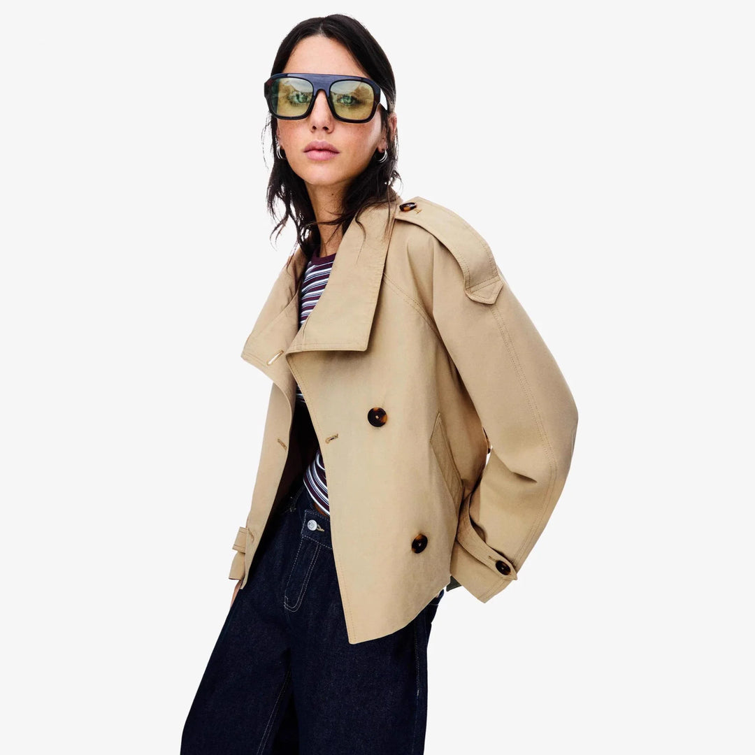 Woman wearing oversized beige trench coat, striped top, dark jeans, and large tinted sunglasses