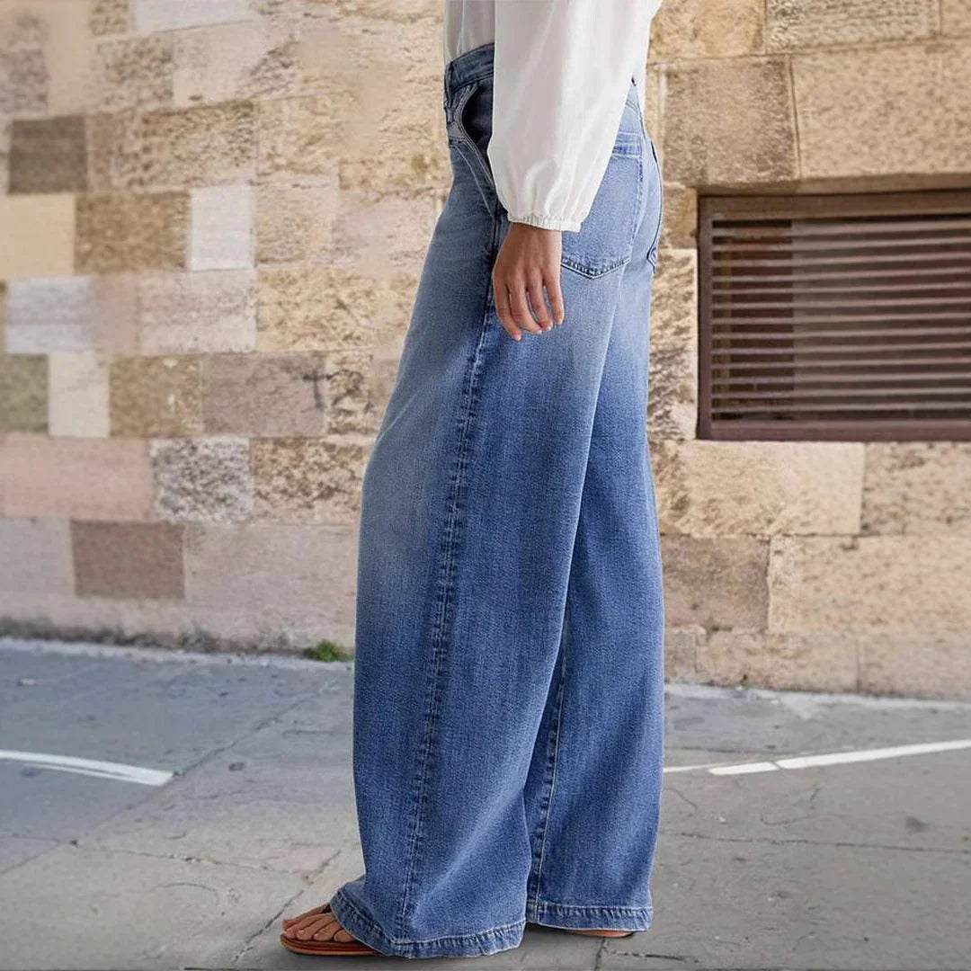 Woman wearing wide-leg blue denim jeans and white long-sleeve top standing on sidewalk