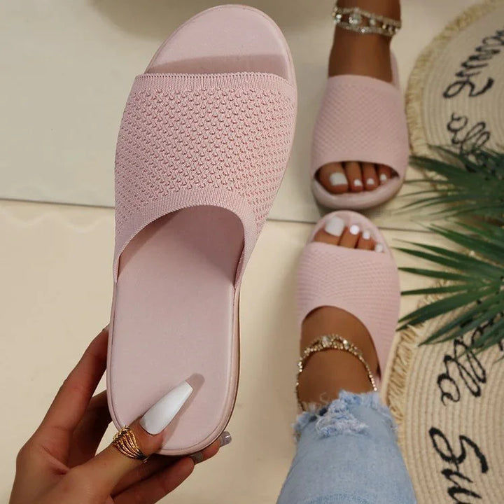 Light pink textured slide sandals worn on feet with white pedicure and denim jeans on beige floor