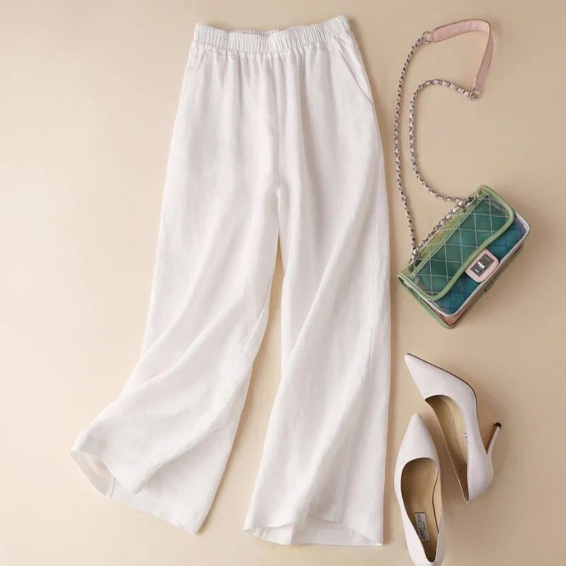 White wide-leg pants with elastic waistband, white pointed-toe heels, and green quilted crossbody bag on beige background
