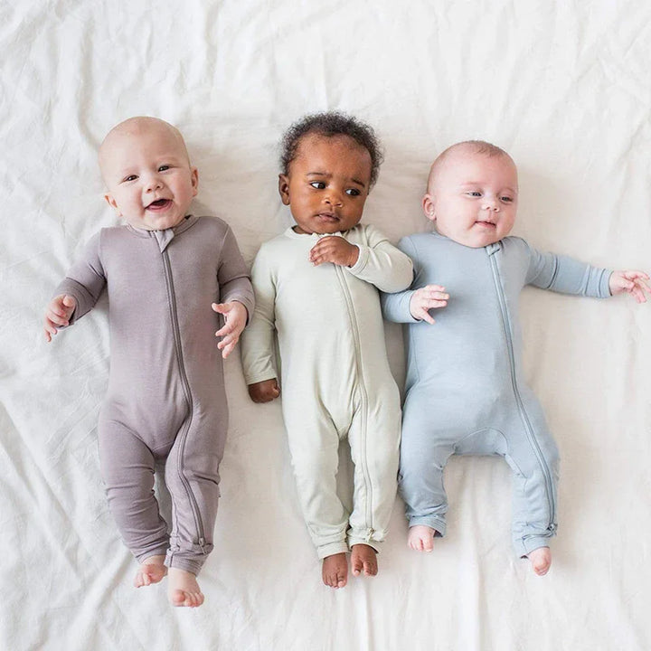 Three diverse babies lying on white sheet wearing pastel colored zipper onesies in gray, cream, and blue