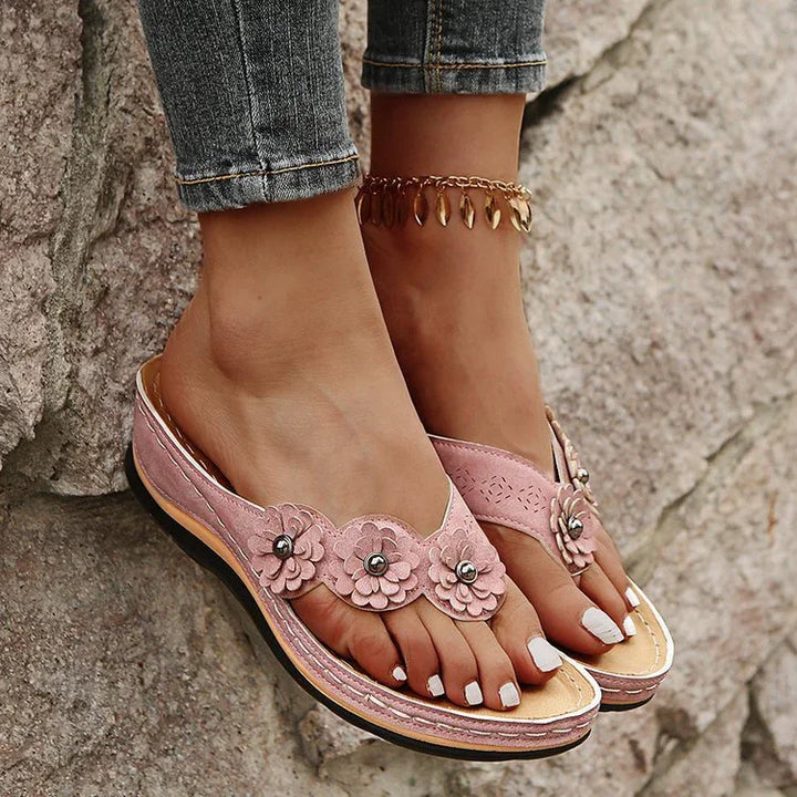Women's feet wearing pink floral leather sandals with white nail polish and gold anklet against textured stone background