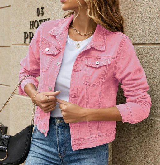 Woman wearing pink denim jacket, white t-shirt, and blue jeans, holding black handbag against stone wall