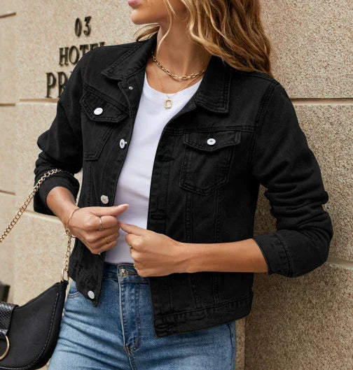 Woman wearing black denim jacket over white shirt with blue jeans, posing by stone wall