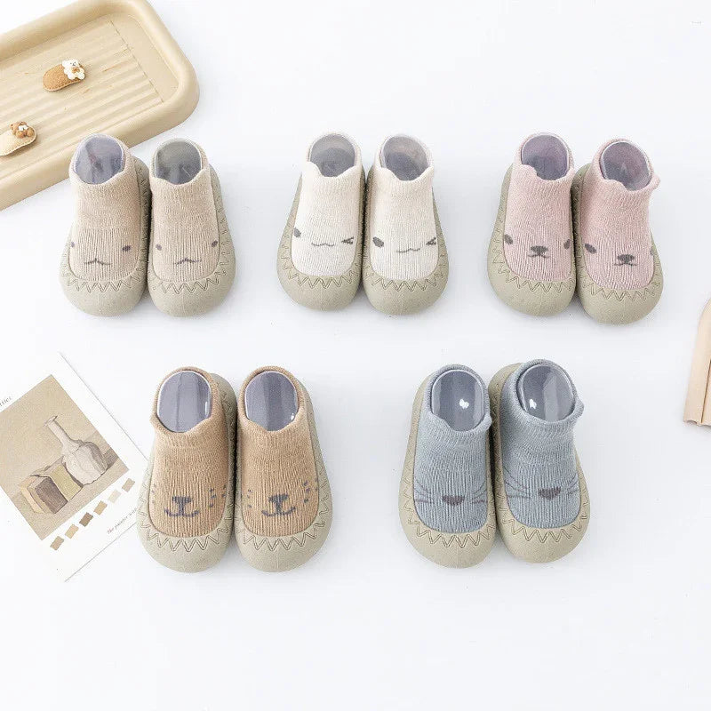 Five pairs of soft baby shoes with animal faces in beige, white, pink, brown, and gray on white background