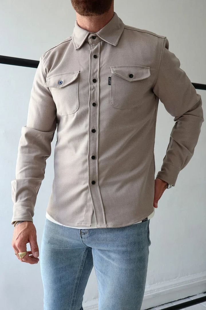 Man wearing beige button-up shirt with chest pockets and light blue jeans in a minimalist indoor setting