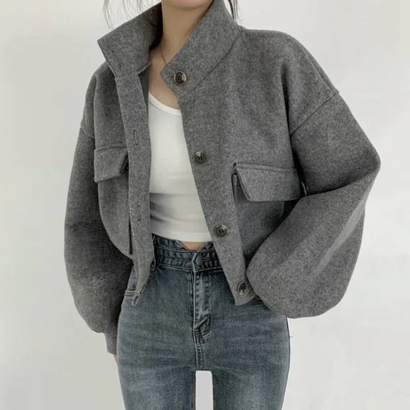 Woman wearing a cropped grey wool jacket with oversized sleeves and front pockets over white crop top and high-waisted jeans