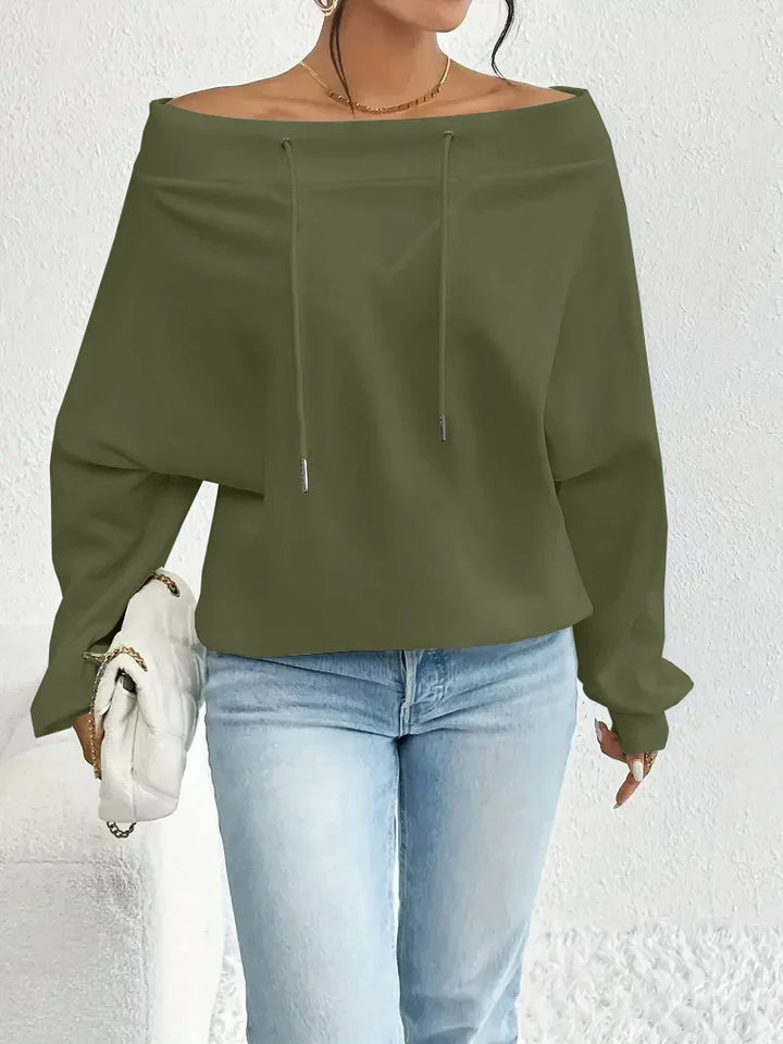 Woman wearing olive off-shoulder sweatshirt with drawstrings and light blue jeans holding white quilted bag
