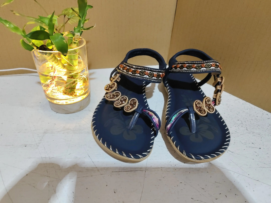Blue floral women's sandals with decorative beads on a white surface next to a lit glass vase with green plant