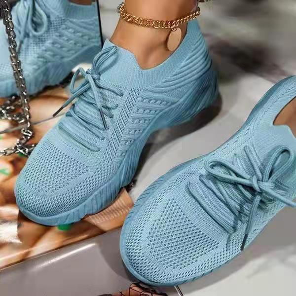 Light blue knitted mesh sneakers with laces and thick soles, worn with a gold anklet on a reflective surface