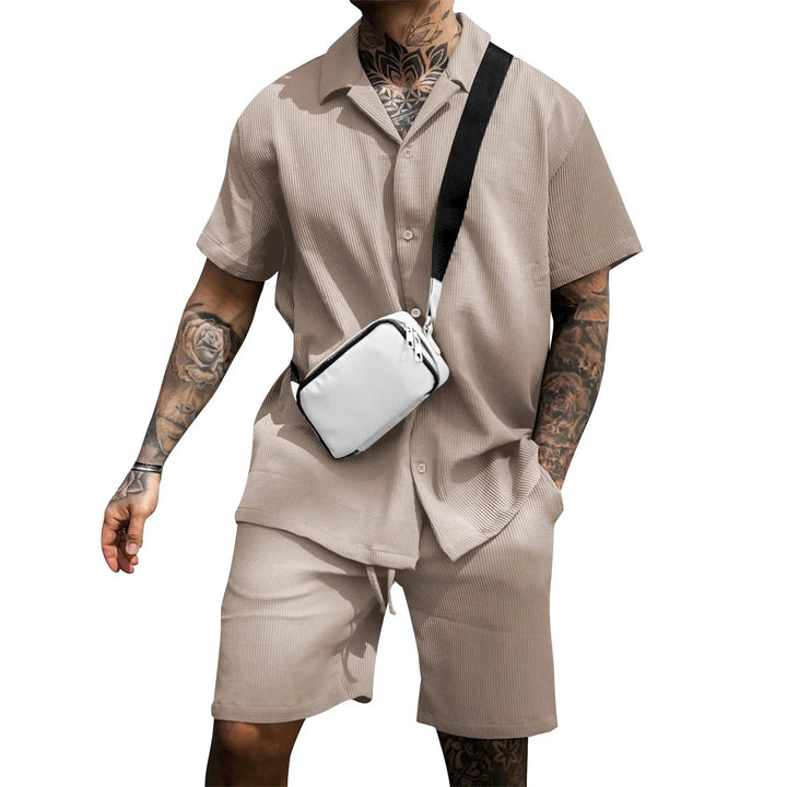 Man wearing beige short-sleeve shirt and shorts set with white crossbody bag and tattooed arms