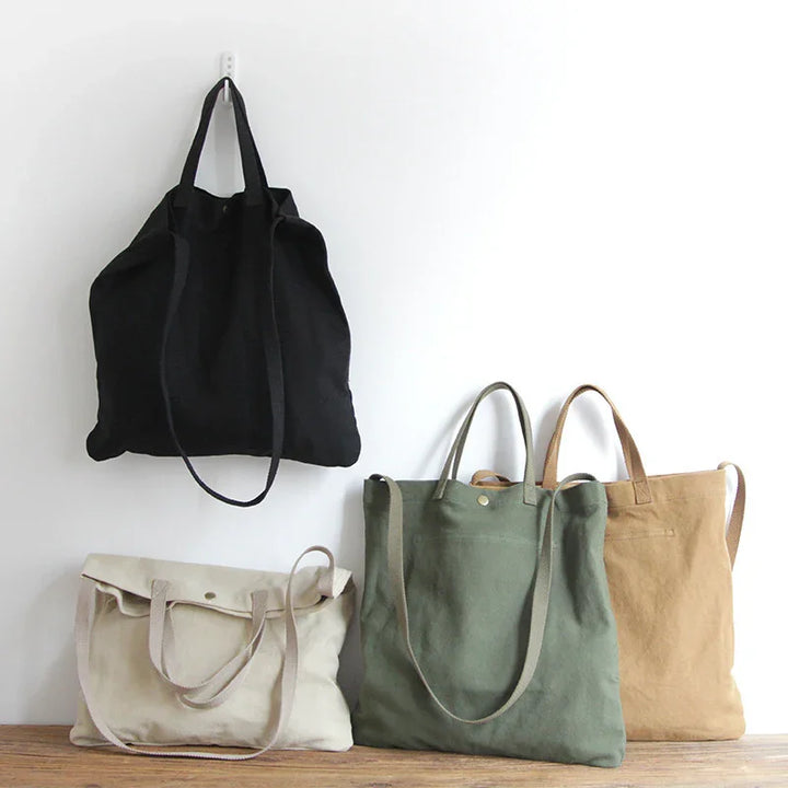 Four canvas tote bags in black, beige, green, and light brown displayed on wall and wooden surface