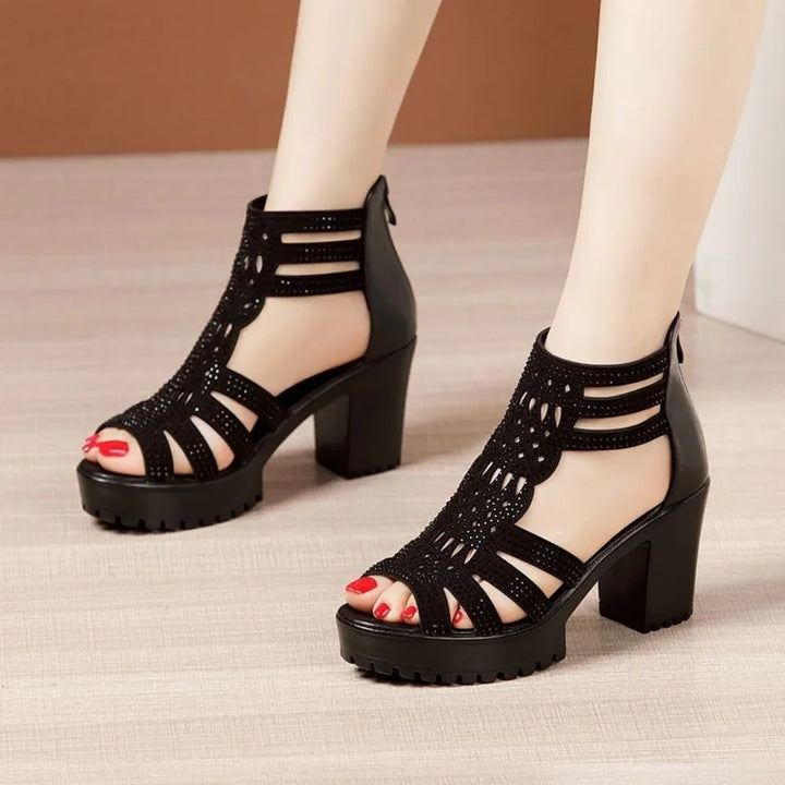 Black high-heel platform sandals with intricate cutout design worn on feet with red nail polish