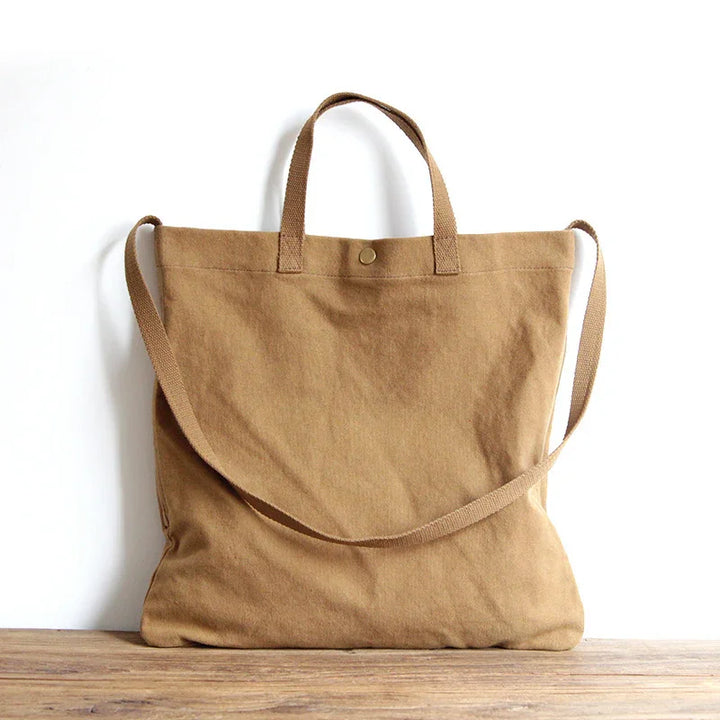 Brown canvas tote bag with short handles and a long shoulder strap on wooden surface