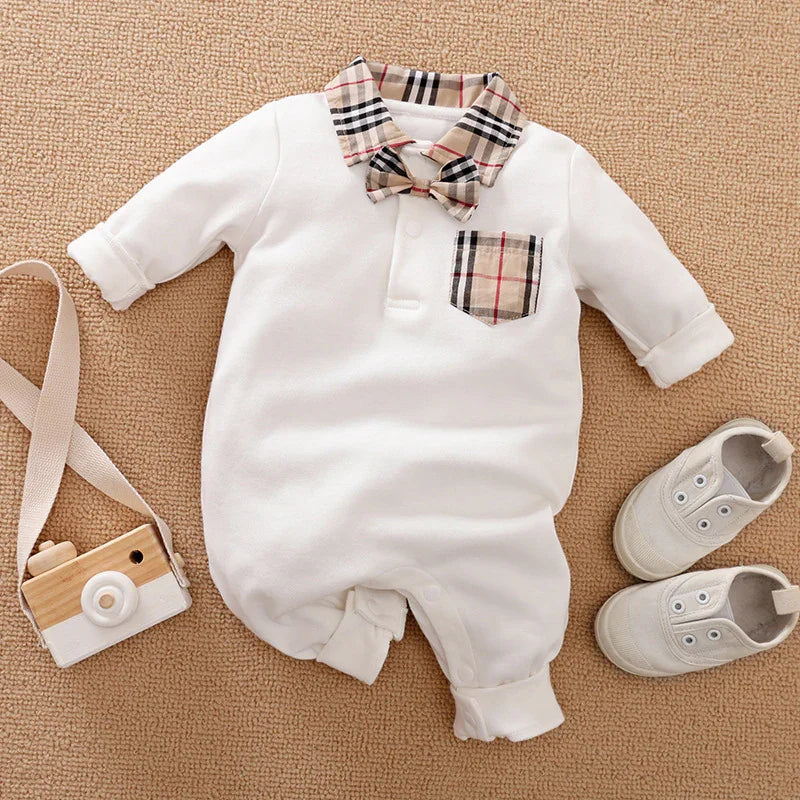 White baby jumpsuit with plaid collar, bow tie, and pocket, paired with white baby shoes and wooden toy camera