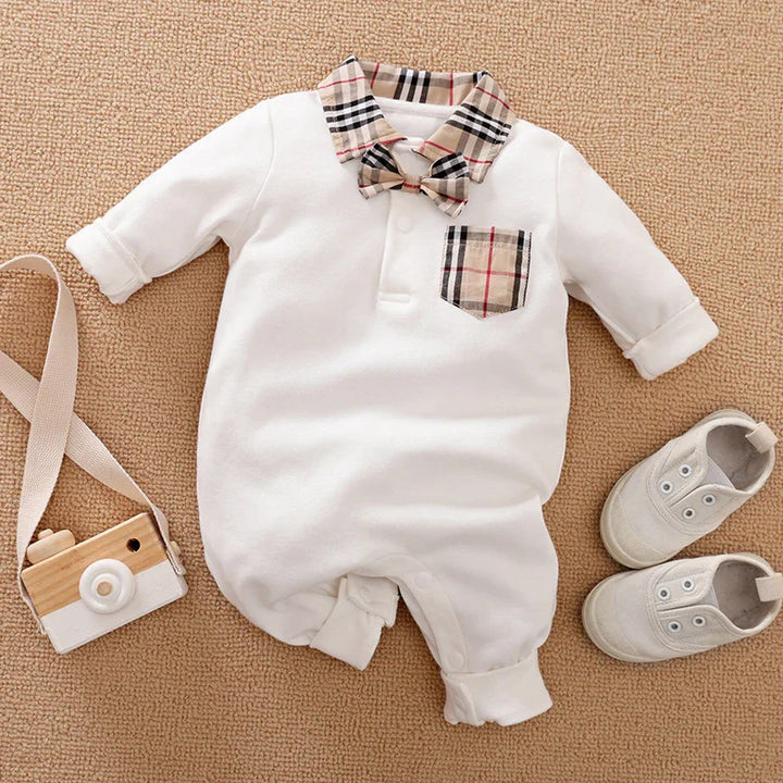 White baby jumpsuit with plaid collar, bow tie, and pocket, paired with white baby shoes and wooden toy camera