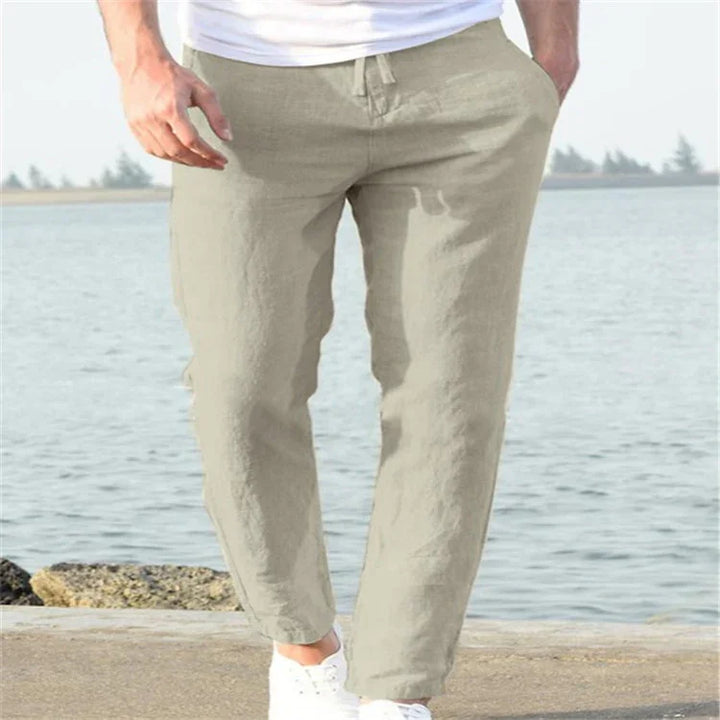 Man wearing beige linen pants and white sneakers standing near waterfront on concrete surface