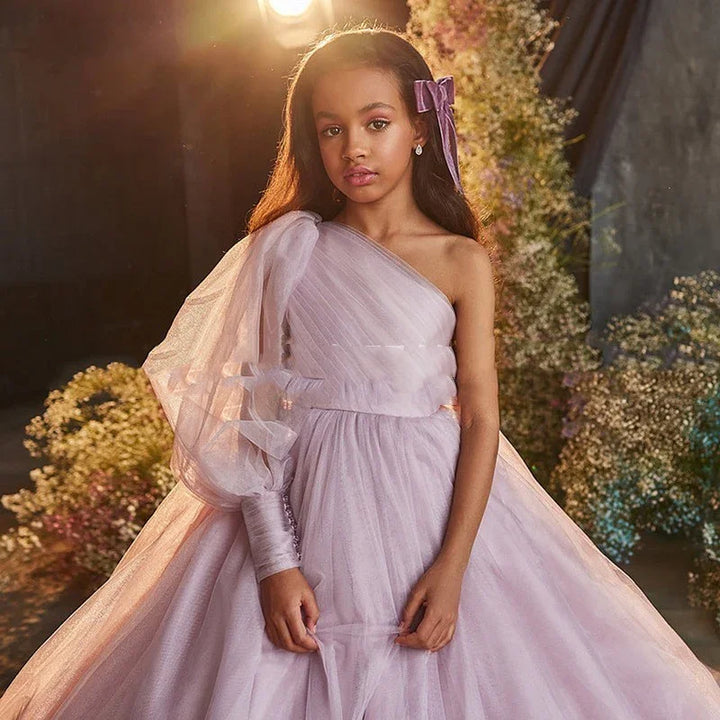 Young girl in elegant lilac one-shoulder tulle dress with bow in hair, floral backdrop, soft lighting