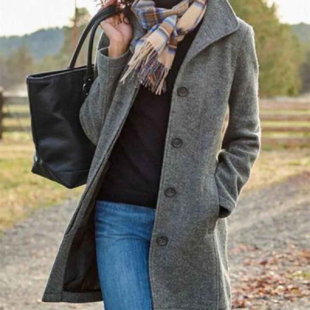Woman wearing grey buttoned coat, plaid scarf, blue jeans, carrying black handbag outdoors