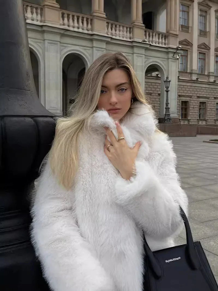 Blonde woman in white faux fur coat posing outdoors with black handbag in front of historic building
