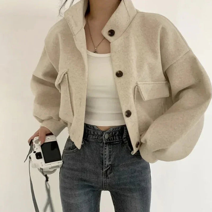Woman wearing beige button-up wool jacket with large pockets, white crop top, and high-waisted black jeans holding camera