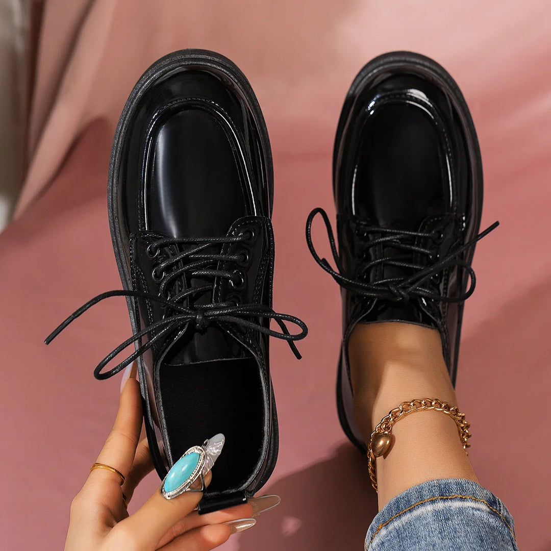 Glossy black leather lace-up shoes worn with jeans and gold anklet on pink background