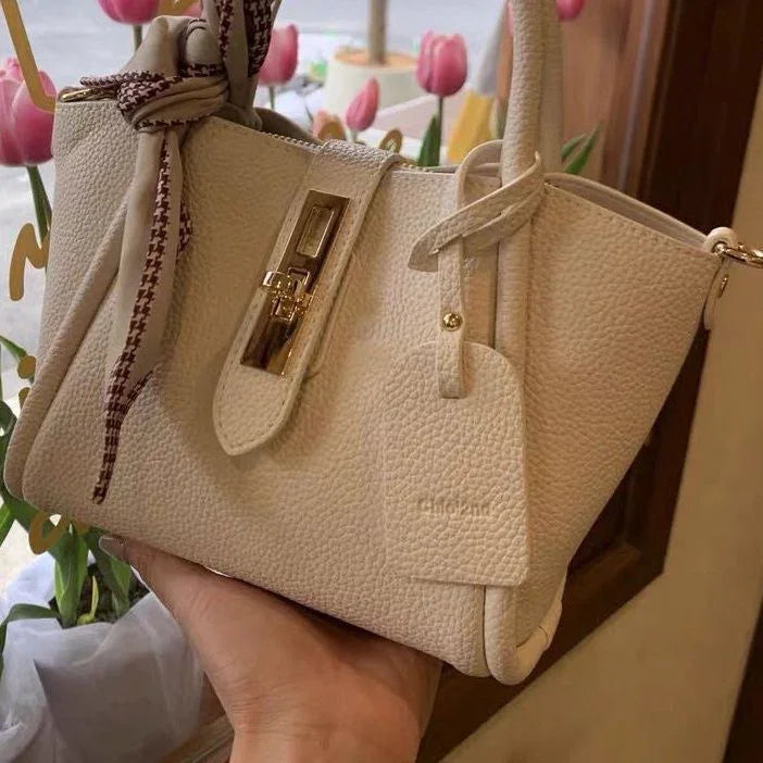 Beige leather handbag with gold-tone hardware and knotted patterned scarf, held in hand near pink tulips