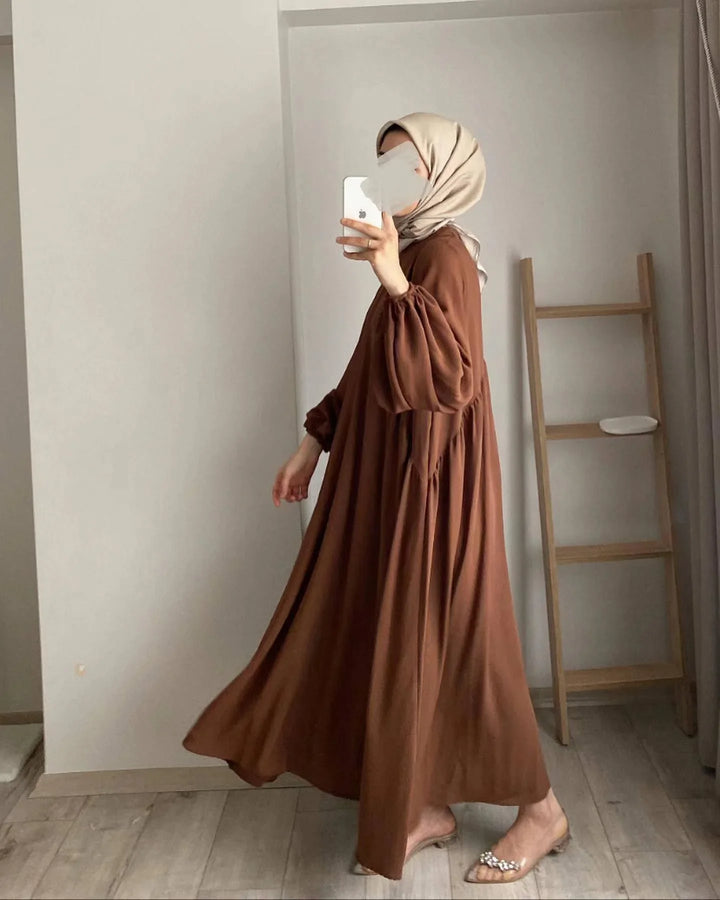 Woman wearing brown long sleeve maxi dress and beige hijab taking mirror selfie indoors