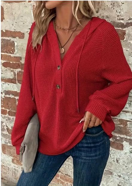 Woman wearing a textured red hoodie with buttons and blue jeans holding a gray clutch against a brick wall