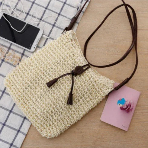 Handwoven beige straw crossbody bag with brown leather strap and button closure on wooden table