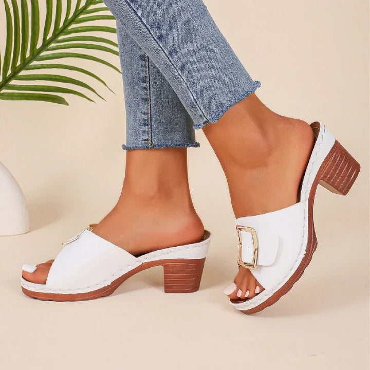 Woman wearing white open-toe mule sandals with gold buckle and chunky wooden heels, paired with frayed hem jeans