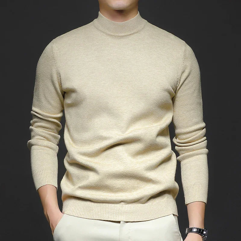 Man wearing beige long sleeve knit sweater with mock neck and light pants against dark background