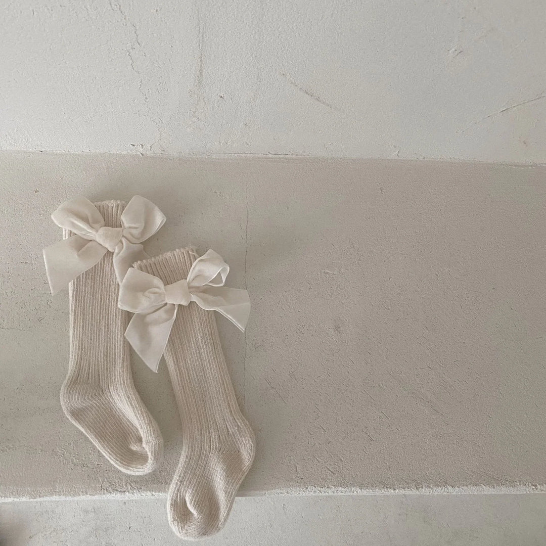 Pair of cream-colored knee-high knitted socks with large white satin bows on a textured beige surface