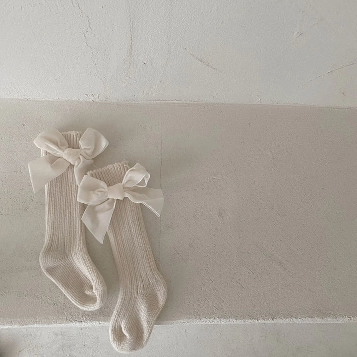 Pair of cream-colored knee-high knitted socks with large white satin bows on a textured beige surface