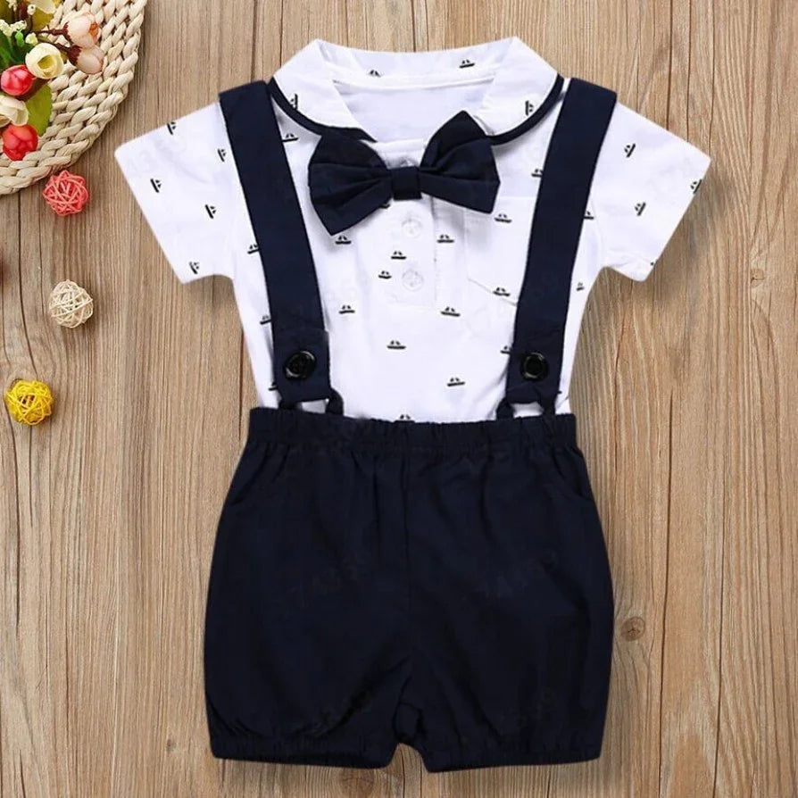 Baby boy formal outfit with white printed shirt, navy bow tie, and navy suspenders shorts on wood