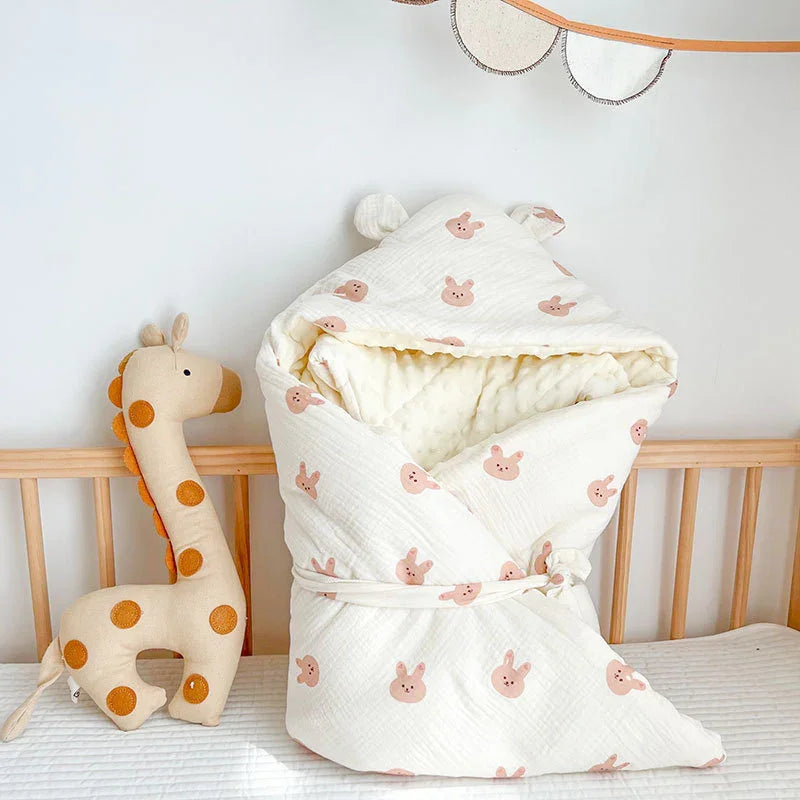 Soft baby swaddle blanket with pink bunny print and ears on hood next to plush giraffe toy in wooden crib