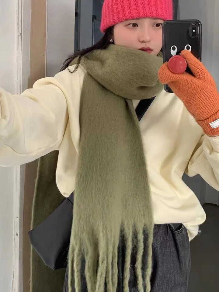 Young woman wearing a red knit beanie, oversized olive green scarf, cream sweater, orange gloves, and holding a black phone with cartoon eyes case