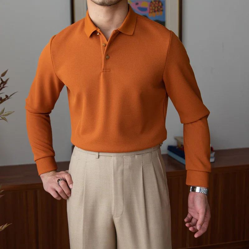 Man wearing an orange long-sleeve polo shirt paired with beige pleated trousers in a modern indoor setting