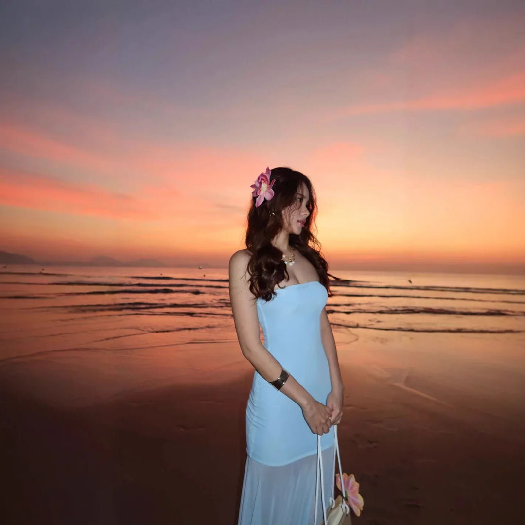 Woman in light blue strapless dress with flower in hair standing on beach at sunset