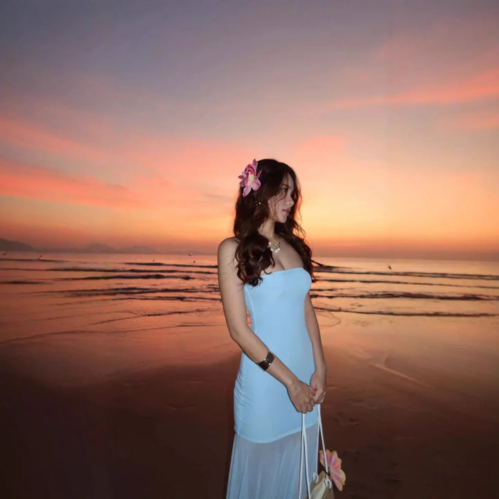 Woman in light blue strapless dress with flower in hair standing on beach at sunset