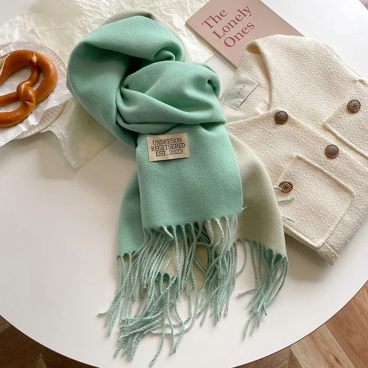 Mint green fringed scarf with Undreson label, cream textured jacket with brown buttons, pretzel on table, and pink book