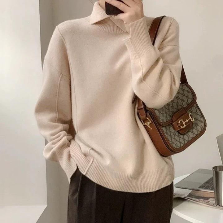 Person wearing beige turtleneck sweater and dark pants, holding brown designer shoulder bag with gold hardware