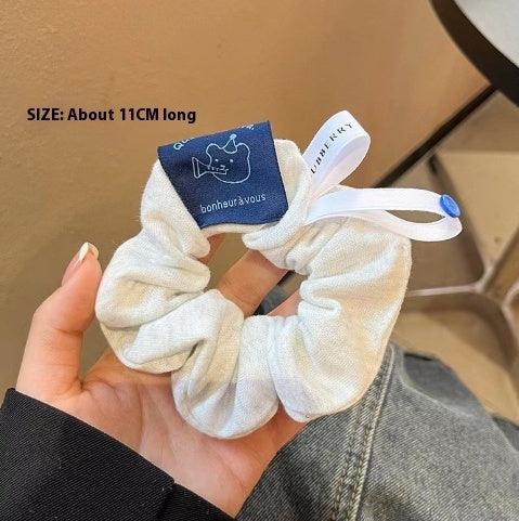 Hand holding white fabric scrunchie with blue tags featuring cartoon dog and text, on beige background