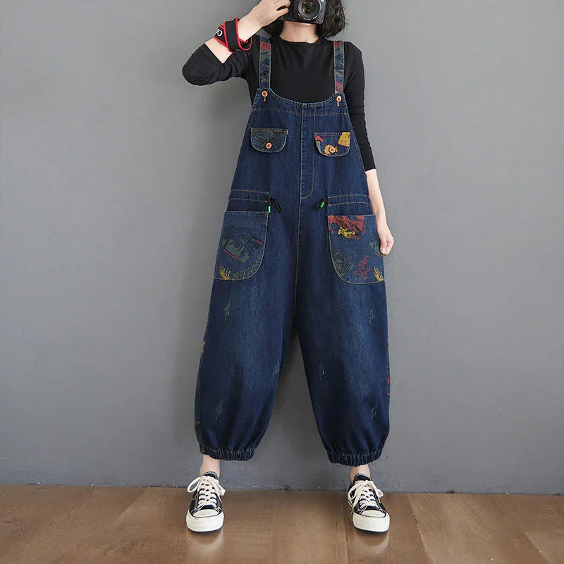 Person wearing loose dark blue denim overalls with large printed pockets over a black long-sleeve shirt and sneakers