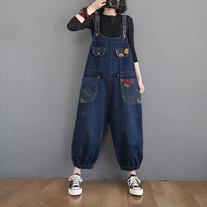 Person wearing loose dark blue denim overalls with large printed pockets over a black long-sleeve shirt and sneakers