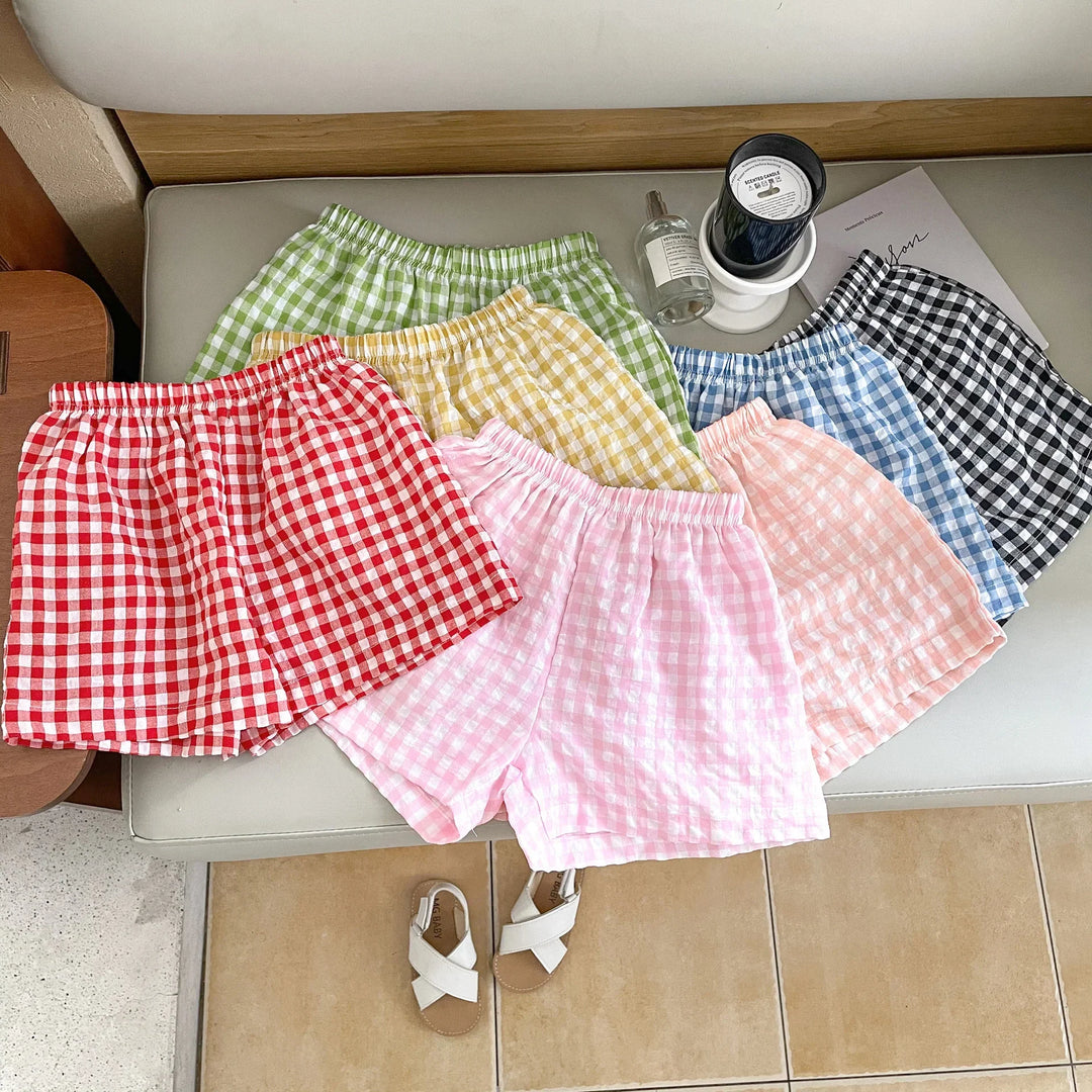 Colorful gingham check cotton shorts in red, green, yellow, pink, peach, blue, and black laid on a bench with white sandals